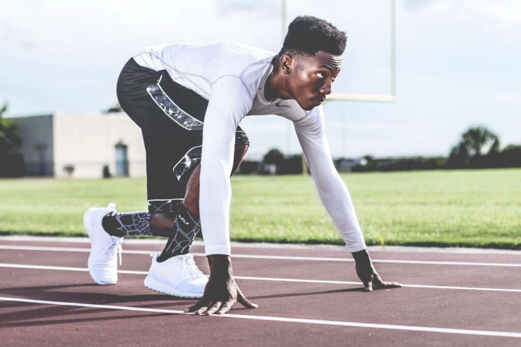 The image shows a young Black man in athletic clothing crouched in a starting position on a track